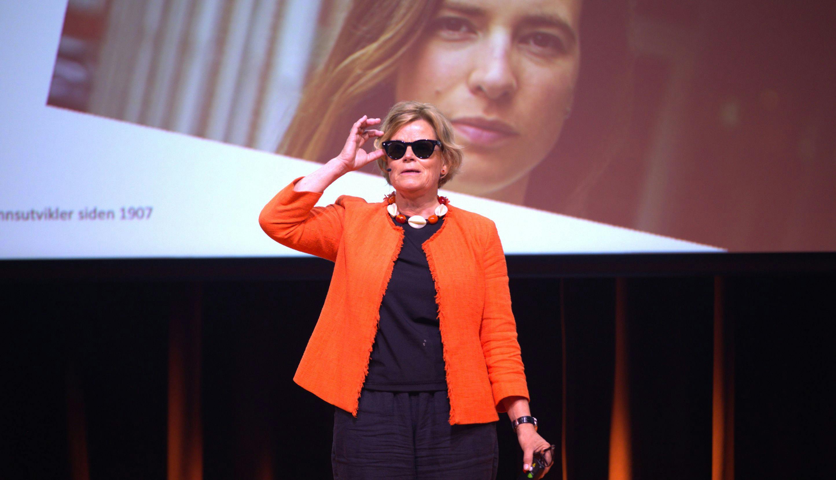 Kvinne med solbriller og oransje jakke på scenen. 