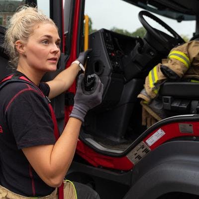 Kvinnelig brannkonstabel står ved brannbil og snakker i walkie talkie. 