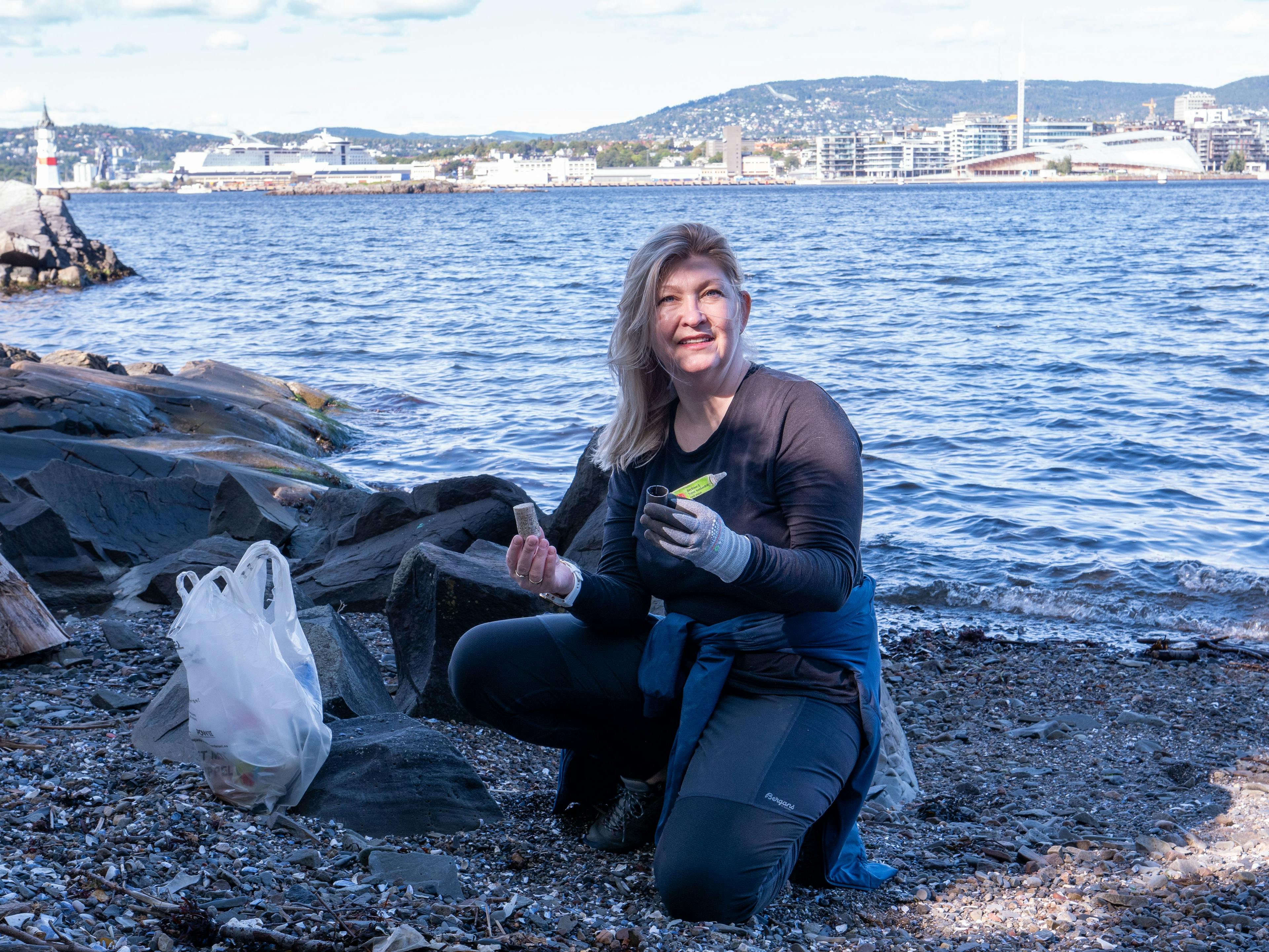 Kvinne som rydder en strand for søppel holder frem noe hun har funnet mens hun smiler mot kamera.