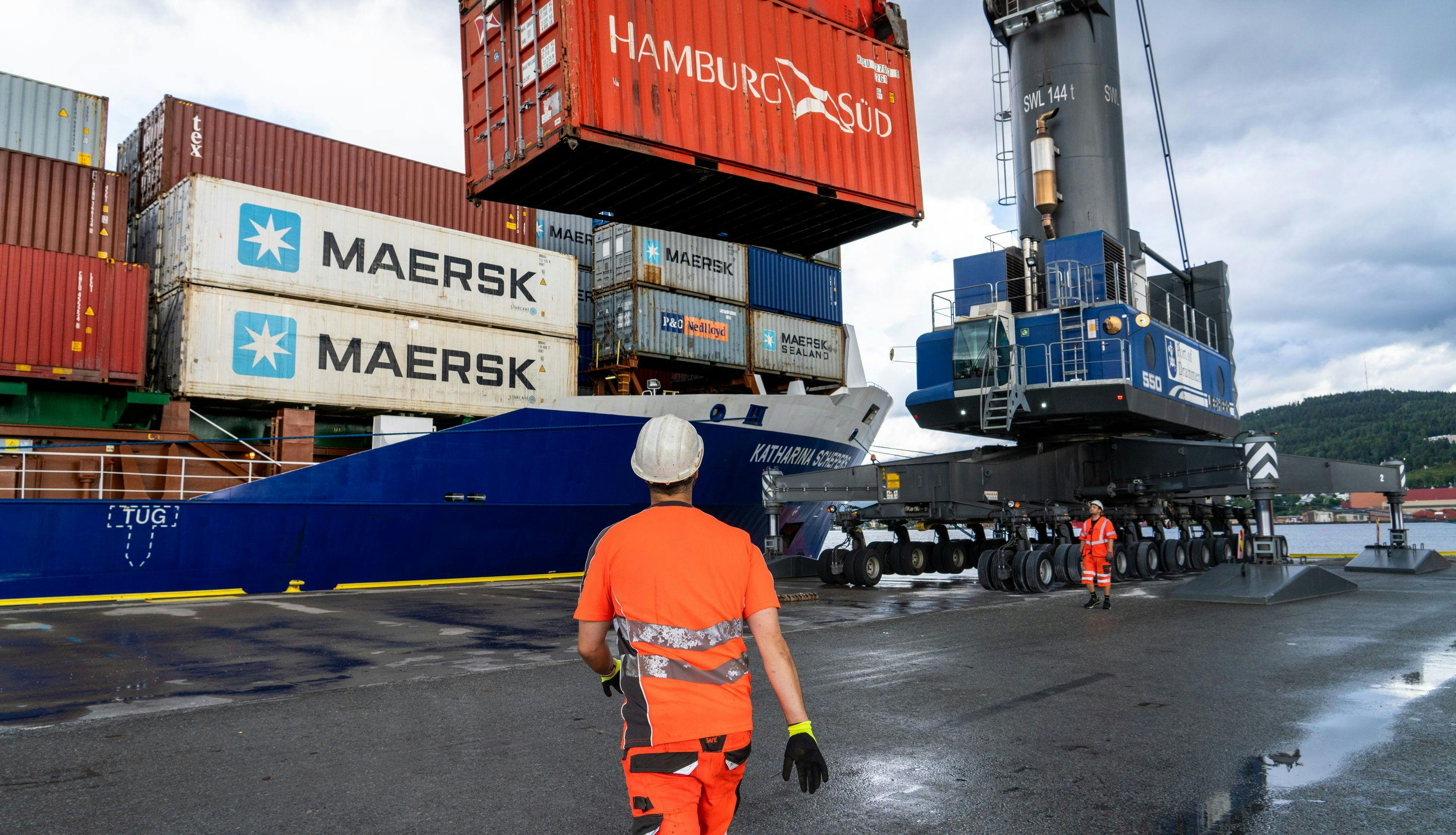 Mann med oransje arbeidsklær og hjelm går mot en containertruck på et havneområde. 