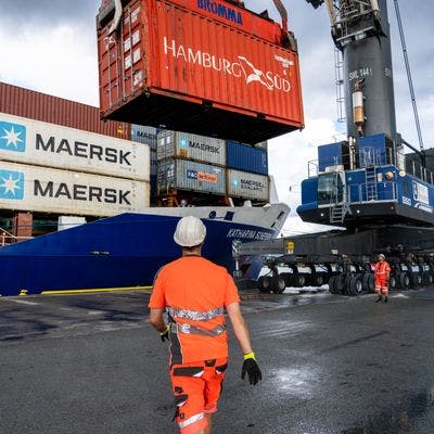 Mann med oransje arbeidsklær og hjelm går mot en containertruck på et havneområde. 