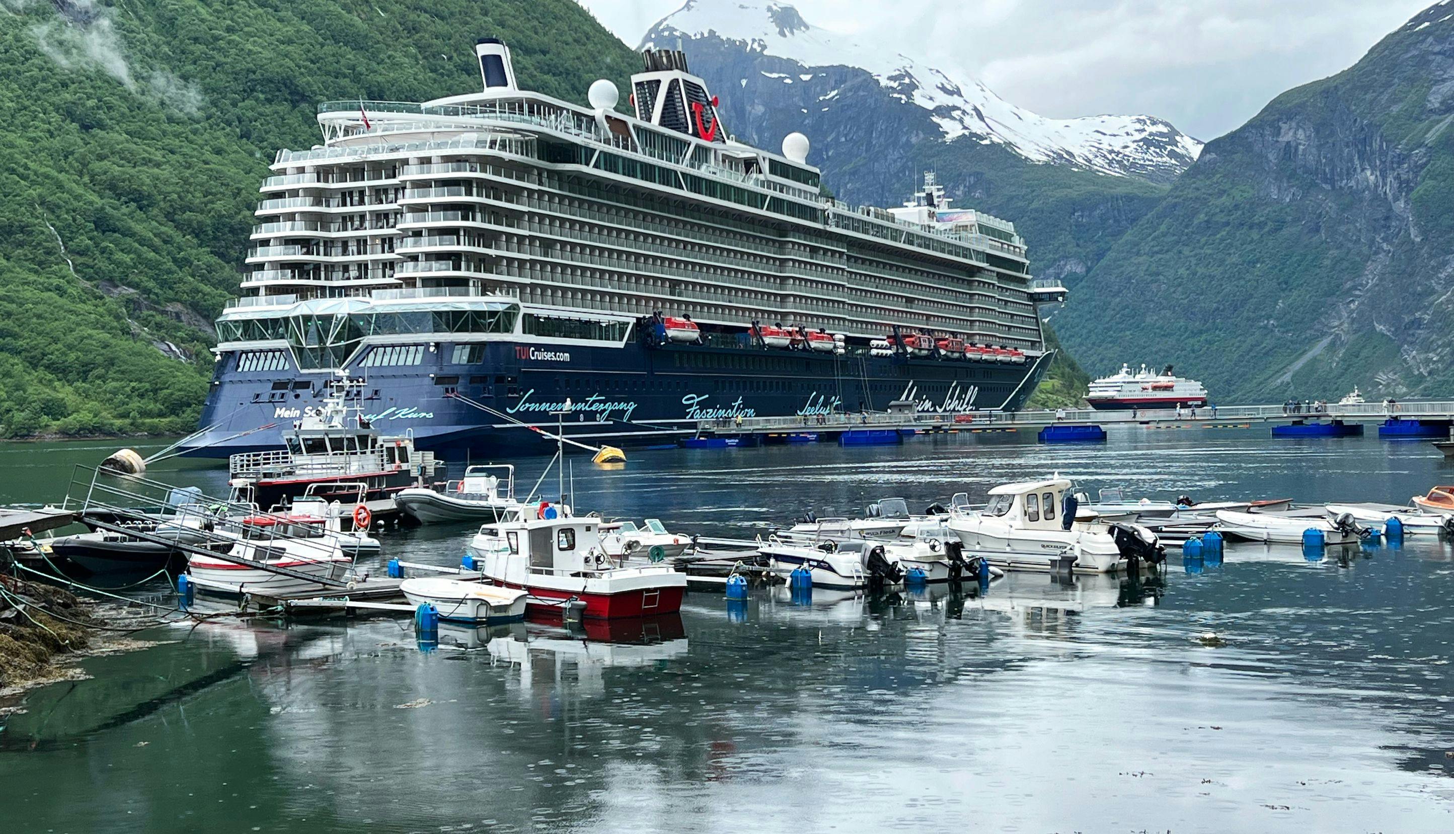 Cruiseskip ligger til kai i en fjord med snødekte fjelltopper i bakgrunnen. 