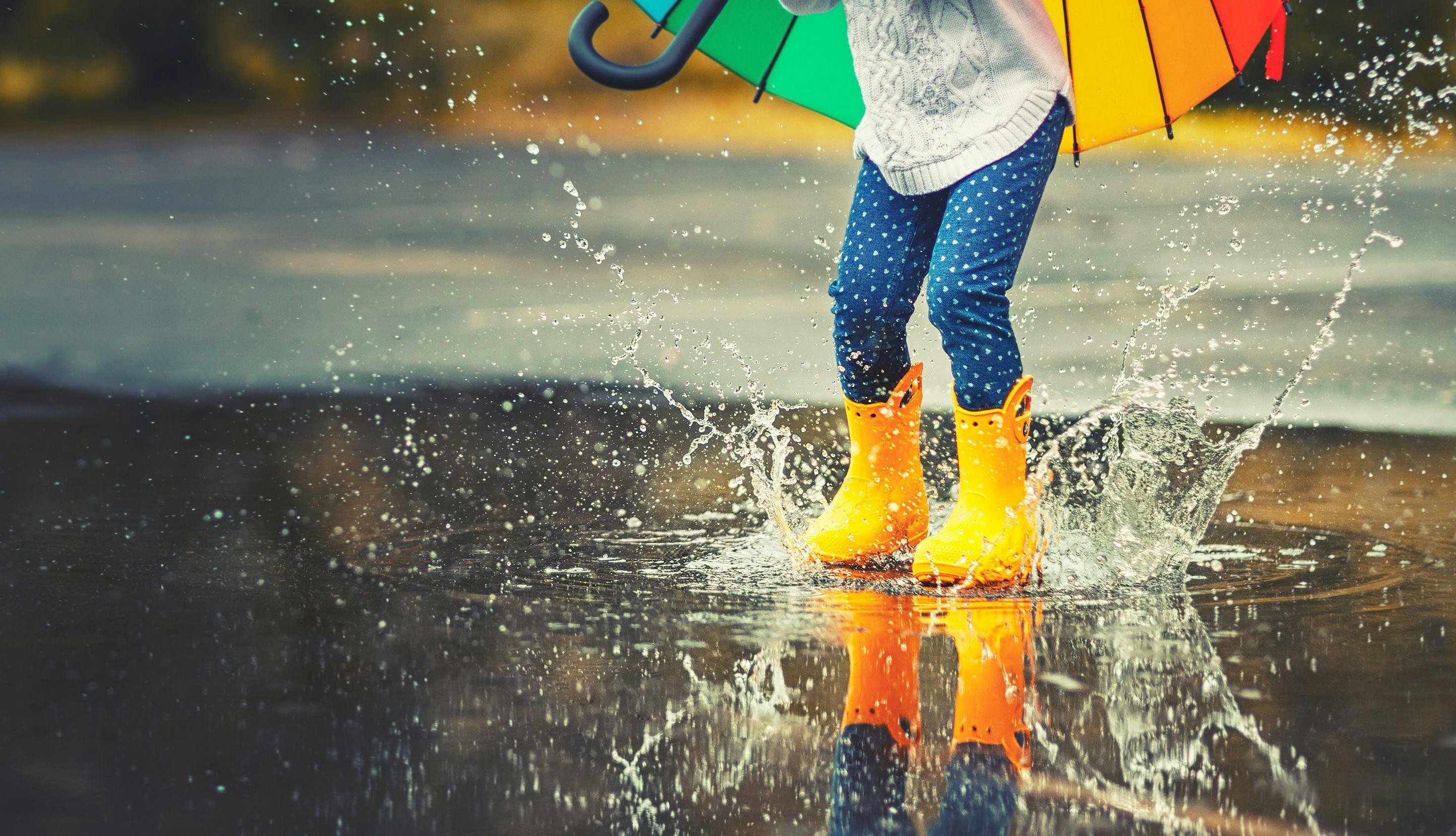 Barn med fargerik paraply og gule støvler hopper i vannpytt så det plasker. 