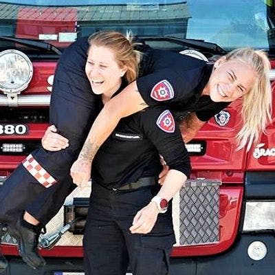 Kvinnelig brannkonstabel bærer en annen kvinnelig brannkonstabel foran brannbil. 